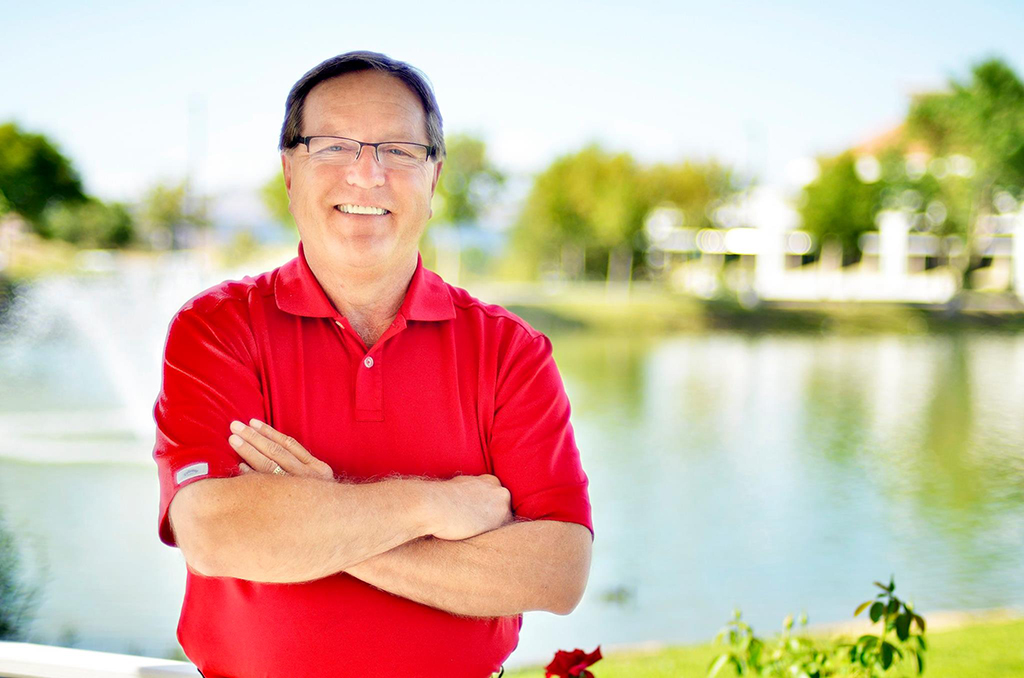Portrait of Bill Greulich smiling in a red polo shirt beside the Victor Valley College campus lake
