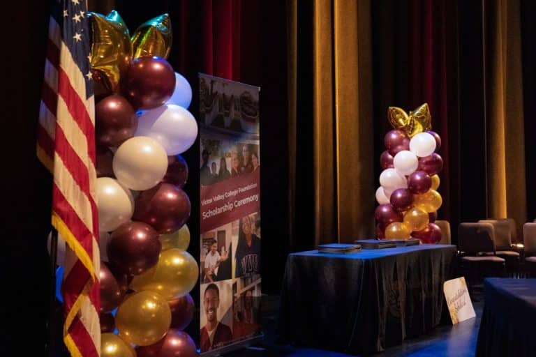 The stage at the Victor Valley College Foundation Scholarship Ceremony, with a United States flag, burgundy, white and gold balloon columns and a Foundation Scholarship Ceremony banner beside the draped awards table