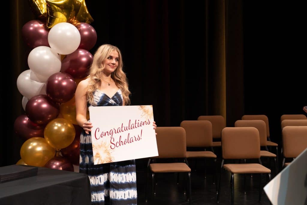 A Victor Valley College Foundation scholarship recipient holds a Congratulations Scholars sign on stage beside burgundy, white and gold balloons