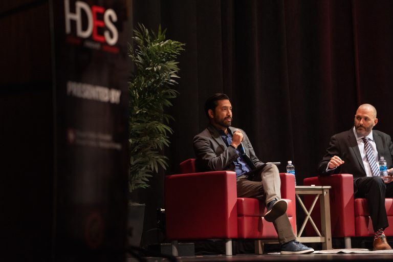 Two panelists seated in red armchairs on stage in front of an HDES banner during a panel discussion