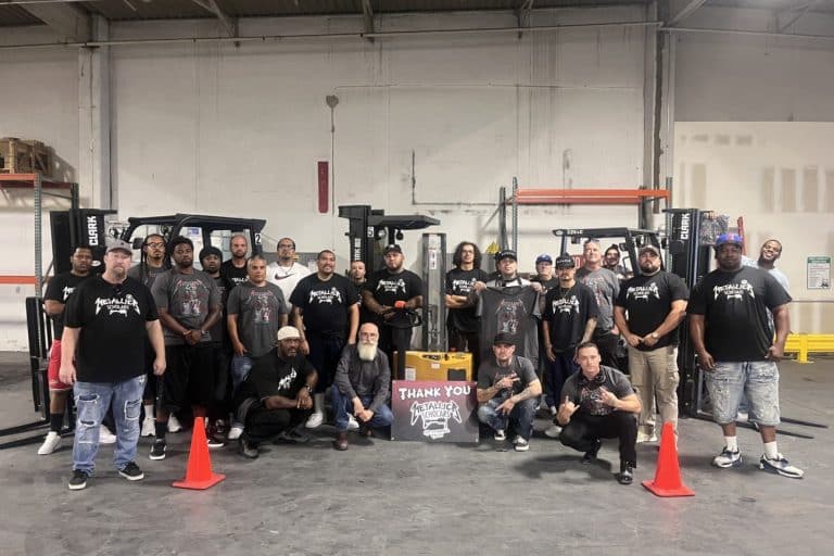 A cohort of Victor Valley College Metallica Scholars in branded shirts standing with forklifts in a training warehouse behind a Thank You Metallica Scholars sign