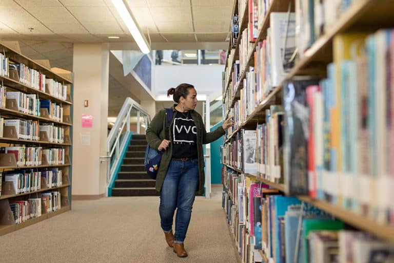 A Victor Valley College student in a VVC shirt browsing books in the campus library