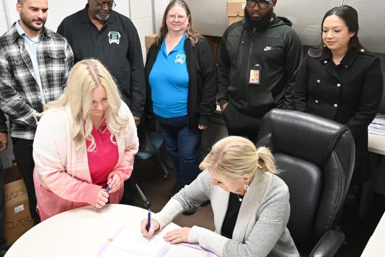Victor Valley Transit Authority and Victor Valley College Foundation representatives gathered around a desk as the paperwork for the donation of four retired buses is signed